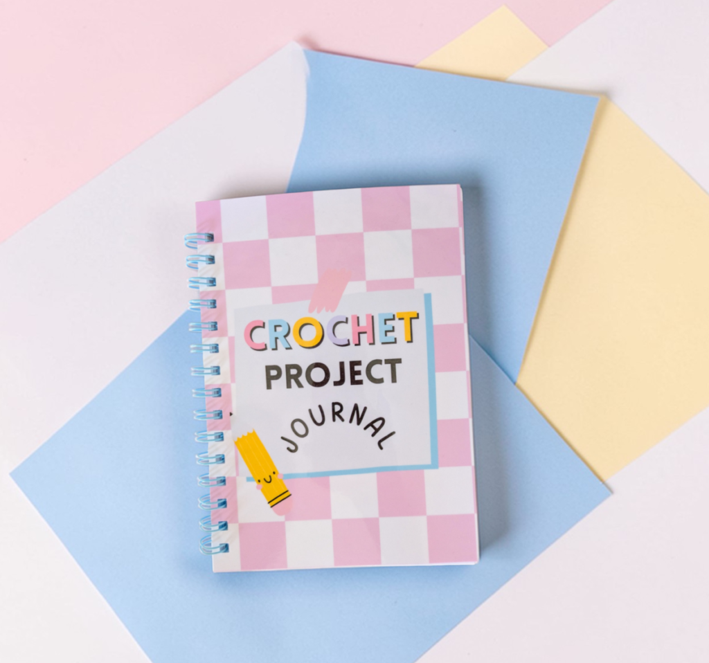 Un carnet à spirales avec une couverture à carreaux roses et blancs sur une surface bleue. Le carnet porte les mots "CROCHET PROJECT JOURNAL" sur la couverture. Un stylo rose est à gauche et à droite du carnet.