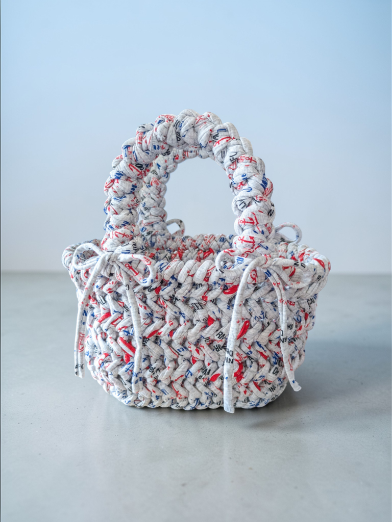 Un petit sac tressé avec une poignée arrondie. Le sac est gris avec un motif de dessins sur le thème nautique rouge et bleu. La poignée et le bord sont faits d'un matériau épais en forme de corde tressée.